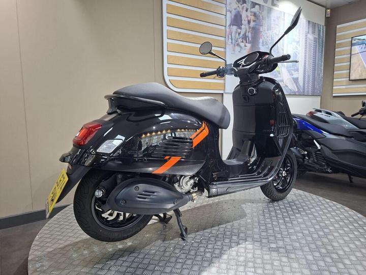 A sleek black scooter with orange accents is displayed in a showroom, showcasing its stylish design and features, including an elevated seat and retro-inspired body shape.