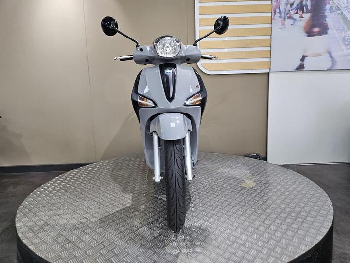 A front view of a sleek, silver scooter displayed on a circular platform, featuring a modern design with headlights and mirrors visible against a neutral background.