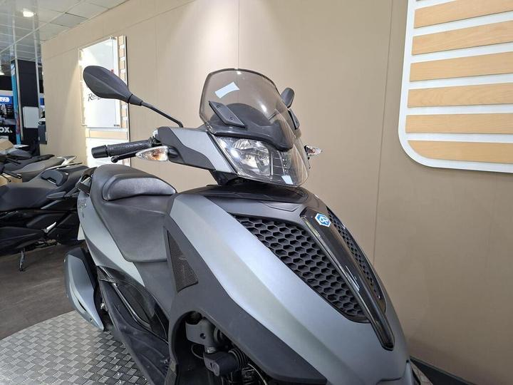 A close-up view of a stylish gray scooter with a sleek design, featuring a transparent windshield and black accents, displayed in an indoor showroom setting.