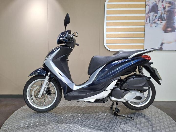 A blue and silver scooter is displayed against a neutral background with wooden accents, showcasing its side profile and sleek design.