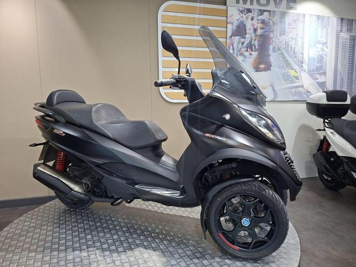 A black three-wheeled scooter displayed in a showroom, featuring a large windshield, comfortable seating, and distinctive alloy wheels. A second scooter in the background is visible, partially obscured. The setting includes modern wall decor and a tiled floor.