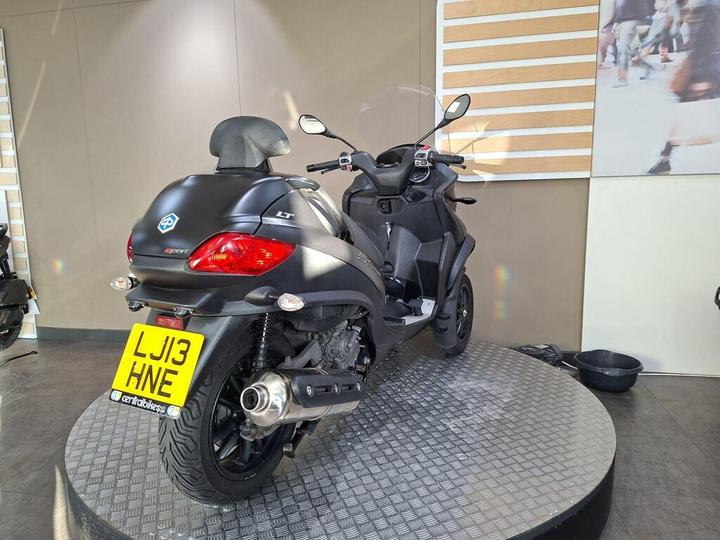 A black scooter displayed on a circular stand, with a license plate reading "LJ13 HNE," set against a neutral backdrop with wooden elements.