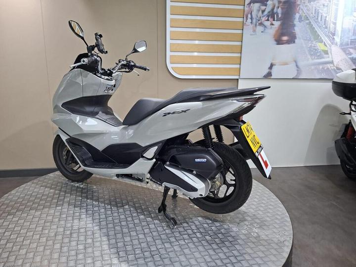 A sleek, gray scooter displayed on a circular metallic platform in a showroom, with a modern backdrop featuring a cityscape. The scooter is viewed from the rear-left angle, showcasing its streamlined design and distinctive tail light.