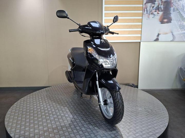 A black scooter displayed on a circular metal platform, with a neutral background featuring a wall with light wooden slats.
