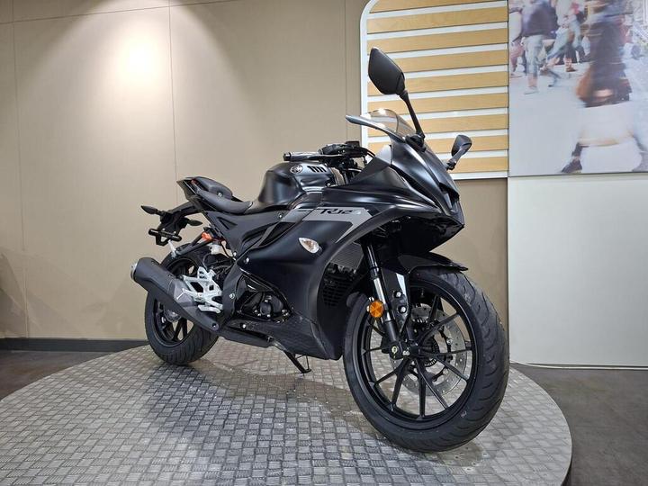 A sleek black sport motorcycle is positioned on a circular platform in a showroom, showcasing its aerodynamic design and modern features. The background features a light-colored wall and blurred figures of people.