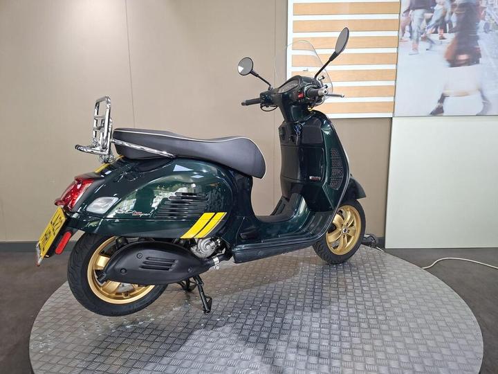 A stylish green scooter with gold wheels displayed on a circular platform in a showroom, featuring a chrome rear rack and a transparent windscreen.