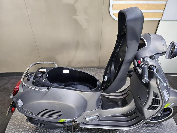 A gray scooter with an open seat, revealing storage space beneath, set against a light-colored wall and patterned floor.
