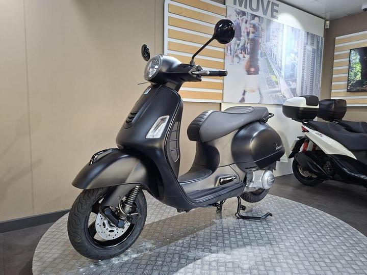 A sleek black scooter with a rounded design is displayed on a textured platform in a showroom, with modern decor and a large graphic backdrop featuring city life.