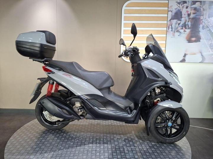 Piaggio MP3 300 Sport 2021 A sleek grey three-wheeled scooter with a large rear storage box, displayed on a textured black surface, set against a neutral wall with a blurred urban scene in the background.