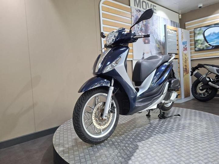 A blue scooter displayed on a circular metallic platform with a beige wall and promotional graphics in the background. The scooter features a sleek design, front headlights, and a comfortable seat.