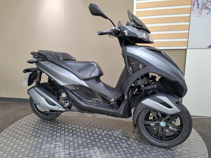 A sleek, modern scooter in a matte gray finish, featuring a streamlined body, dual front wheels, and a comfortable seat, displayed against a neutral background.