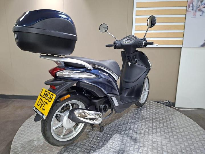 A dark blue scooter with a rear top case, parked on a textured circular platform, with a minimalistic indoor background.
