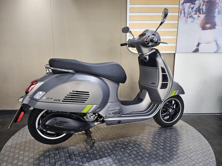 A sleek gray scooter with green accents, displayed on a circular textured platform, set against a neutral background.
