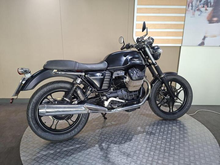 A black motorcycle with a sleek design is displayed on a circular platform, featuring a low-profile seat, chrome exhaust, and modern handlebars, set against a muted background.