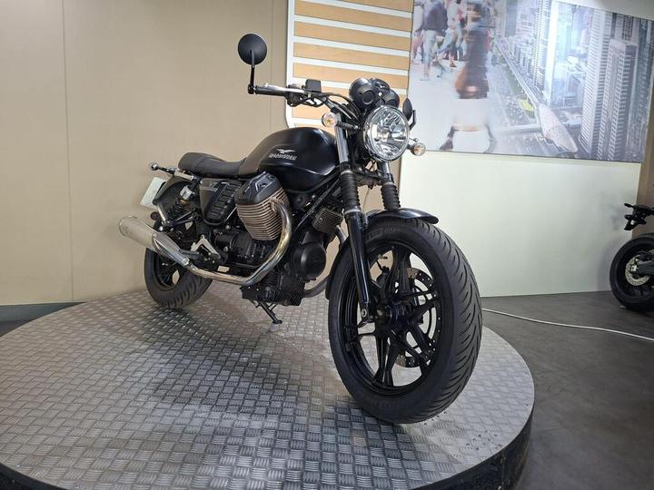 A black motorcycle with a sleek design is displayed on a circular platform in a modern setting, featuring a backdrop of a cityscape. The bike has a prominent front headlight, chrome exhaust, and distinctive bodywork, while the floor has a textured pattern.