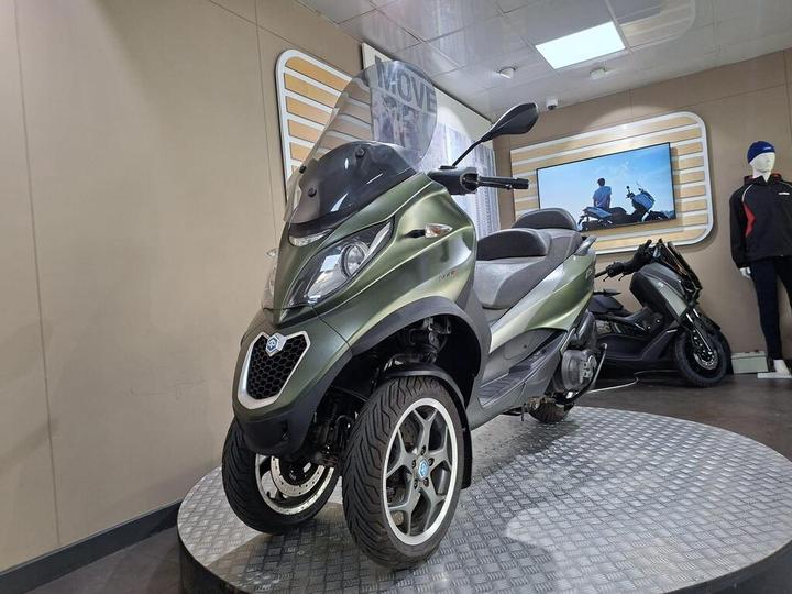 A green three-wheeled scooter displayed on a circular platform in a showroom, with a person wearing a black and red jacket in the background and a television screen showing another scooter.
