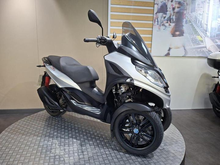 A silver three-wheeled scooter displayed on a patterned platform against a neutral background.