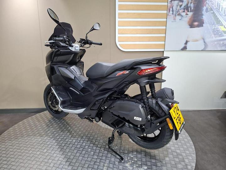 A sleek, black scooter is positioned on a textured display surface, showcasing its profile from the rear-left angle. Features include a high windshield, modern design elements, and reflective rear lights, with a registration plate visible. A blurred urban background adds context.