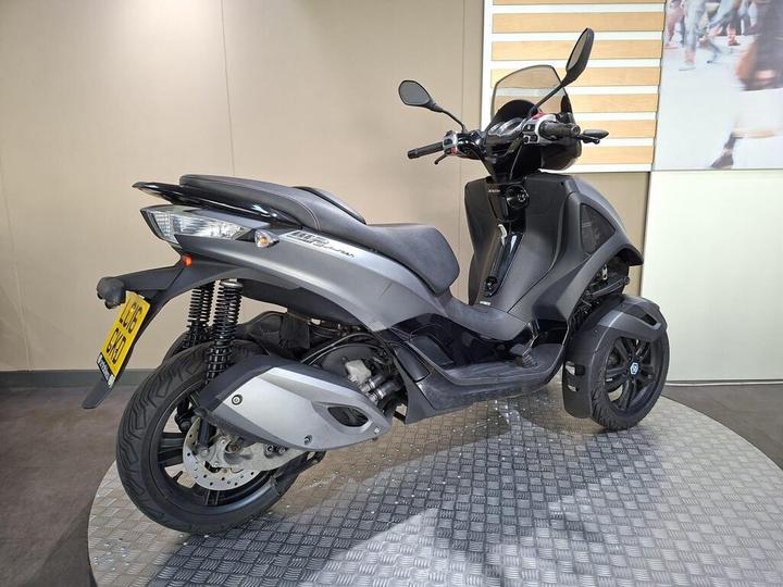 A sleek, modern three-wheeled scooter is displayed on a textured surface, featuring a gray body, black seat, and ergonomic handlebars. The background is neutral, highlighting the scooter's design.