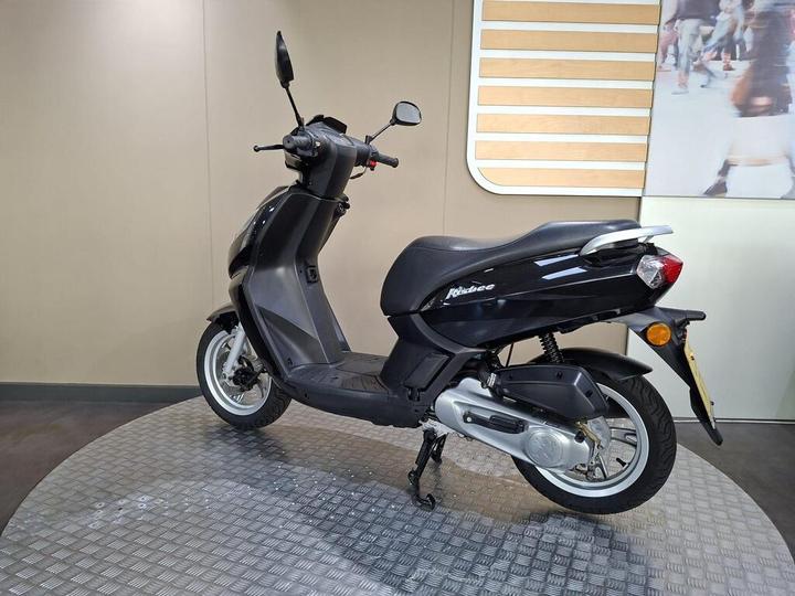A black scooter is displayed on a circular platform, featuring a sleek design with a low seat and large wheels. The background is a neutral tone with wooden accents.
