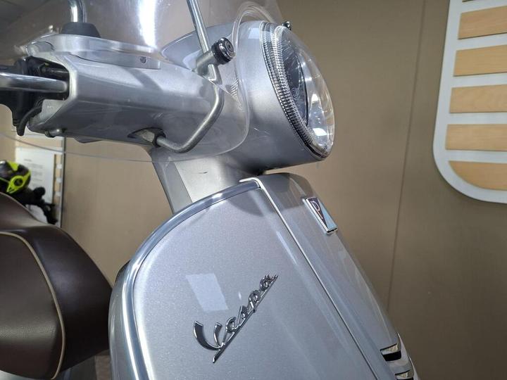 Close-up of a silver Vespa scooter's front, featuring the headlight and iconic logo, with a blurred background of a showroom.