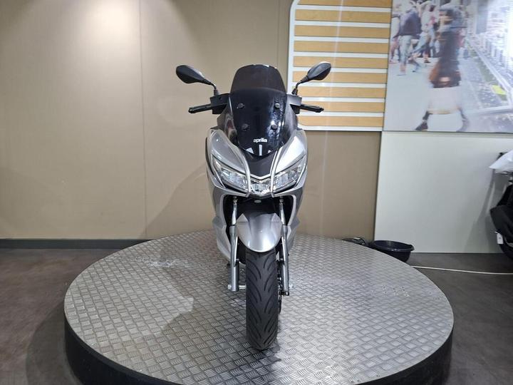 A silver motorcycle displayed on a rotating platform against a neutral background, featuring a modern design with a prominent front view, headlights, and mirrors.