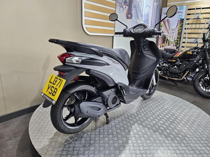A gray scooter with a sleek design is displayed on a circular platform in a showroom. The scooter features a black seat, rearview mirrors, and distinctive rear lights. A motorcycle is partially visible in the background. The wall behind features a light wood paneling design.