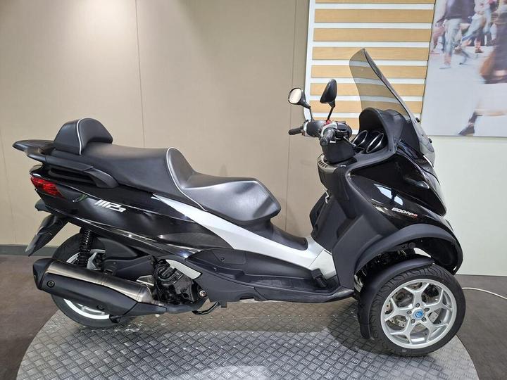 A sleek black and silver three-wheeled scooter displayed in an indoor showroom setting, showcasing its modern design and features.
