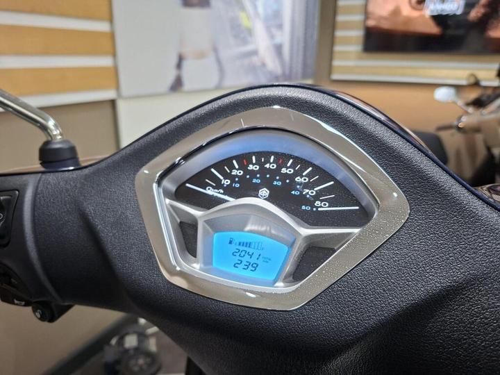 Close-up of a scooter's dashboard displaying speedometer and odometer readings; speedometer indicates 0 km/h while the odometer shows 204.1 km. The background is blurry with some interior design elements visible.