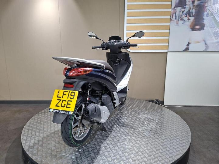 A black and silver scooter is positioned on a round metal platform, showing its rear and profile. The scooter's license plate reads "LF19 ZGE." The backdrop features a light-colored wall and a large image of people walking.