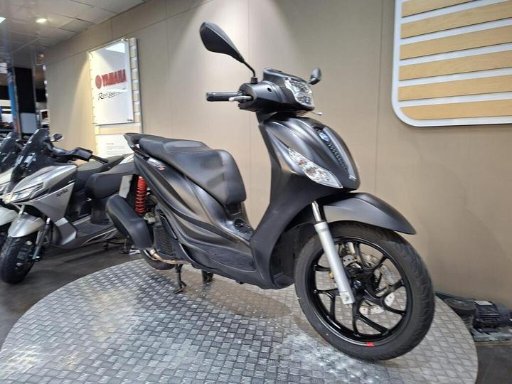 A sleek black scooter displayed on a circular platform with a checkered pattern, featuring a modern design, visible headlights, and a prominent logo in the background.