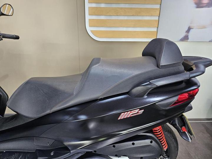 A close-up of a sleek, black scooter seat featuring modern design elements, with red suspension visible at the rear and a minimalistic background.