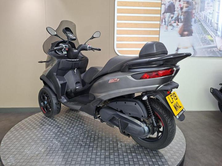 A grey three-wheeled scooter, model MP3 500, positioned on a circular platform with a patterned surface. The scooter features a rear view with distinctive red accents and a license plate displaying "LF19 WKL." The background shows a neutral-colored wall with a blurred city scene.