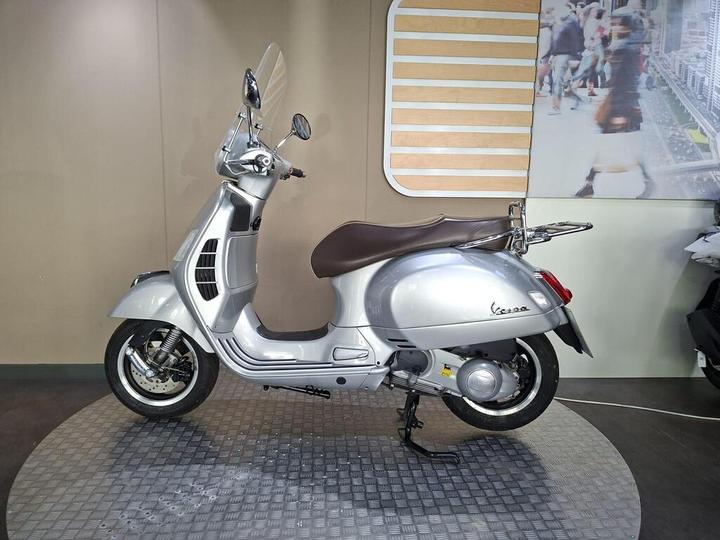 A silver Vespa scooter is displayed on a circular platform against a neutral background, with a modern design featuring a brown seat and protective windscreen.