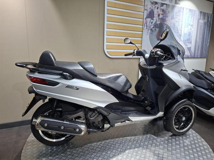 A sleek silver three-wheeled scooter displayed in a showroom, featuring a black seat, rearview mirrors, and modern design elements, set against a neutral background with a cityscape image.