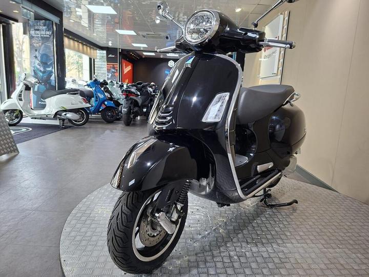 A sleek black scooter displayed in a motorcycle showroom, featuring chrome accents, a round headlight, and a modern design, with several other scooters visible in the background.