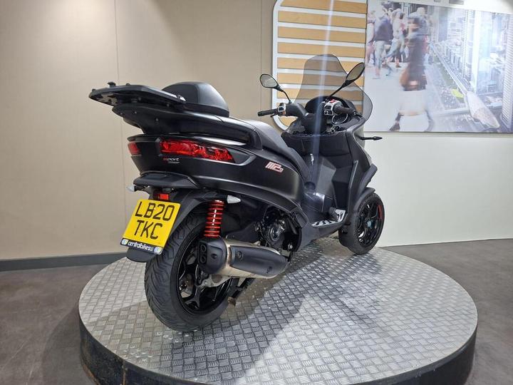 A black three-wheeled scooter displayed on a circular platform, showcasing its rear and side profile, with a UK license plate visible. The background features a digital screen displaying urban imagery.