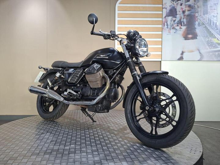 A matte black motorcycle is positioned on a textured circular platform, showcasing its sleek design and chrome exhaust. The background features a light-colored wall with an abstract design, while the right side of the image includes a blurred scene of people walking.