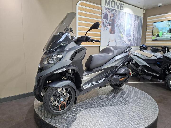 A grey three-wheeled scooter displayed on a circular platform, featuring a large windshield, a sleek design, and located in a showroom with promotional signage in the background.