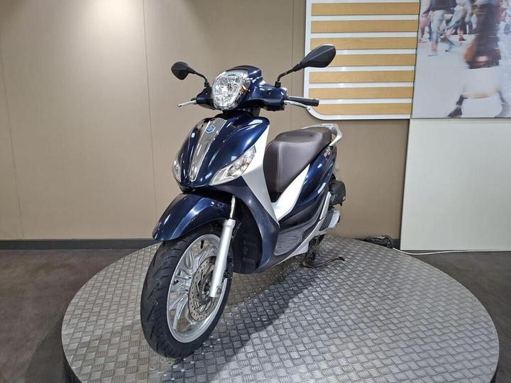 A blue and silver scooter is displayed on a circular metallic platform, with a plain wall in the background.