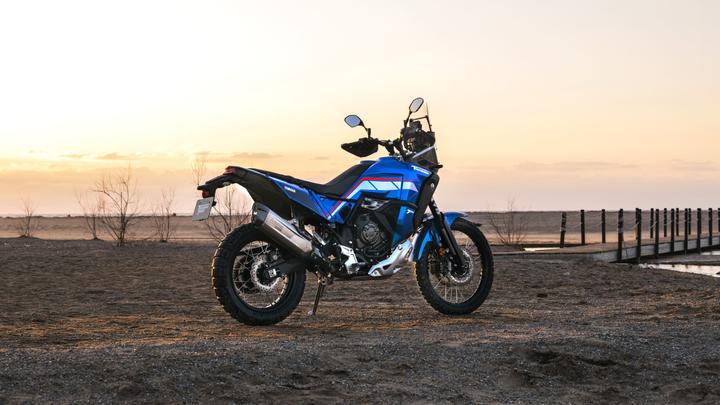 A blue and black adventure motorcycle parked on a sandy terrain during sunset, with a wooden pier in the background.