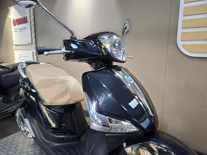 A black scooter with a beige seat and chrome detailing, displayed in a showroom with a beige wall background and Yamaha signage.