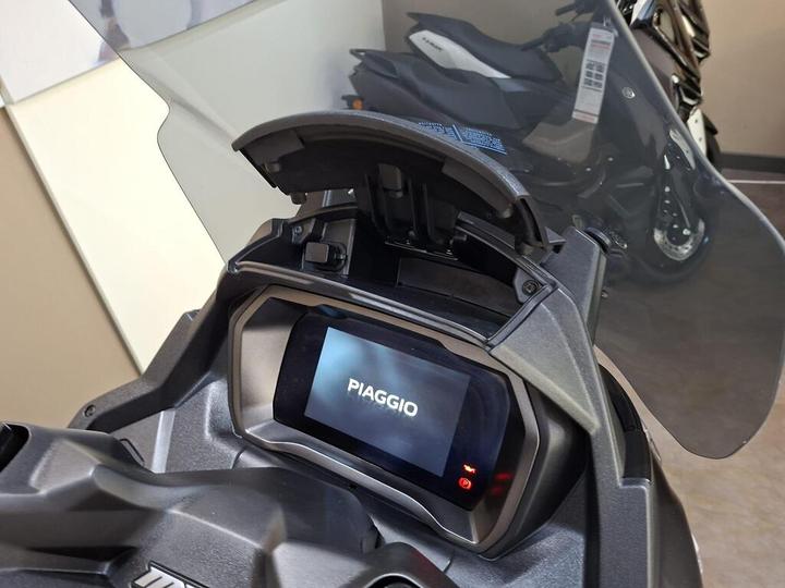 Close-up of a Piaggio scooter's dashboard displaying the brand name on a digital screen, with a partially opened windshield above it and another scooter in the background.