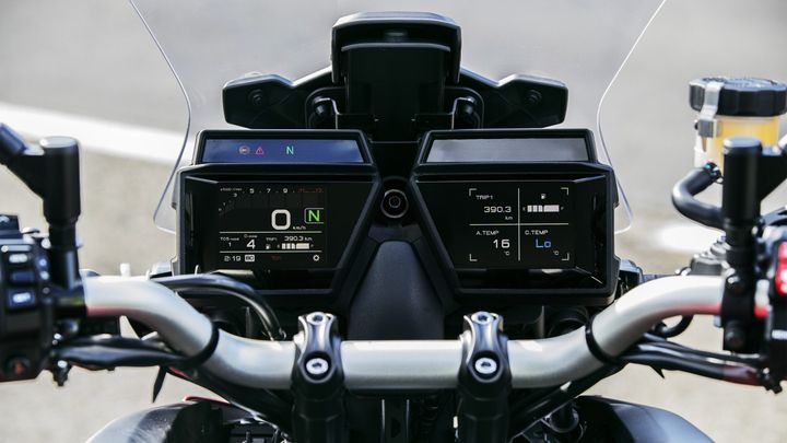 Close-up view of a motorcycle's dashboard displaying digital gauges, including speed (0 km/h), trip distance (390.3 km), temperature readings, and various warning indicators, with handlebars and controls visible.
