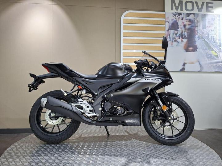 A sleek black motorcycle displayed on a circular metal platform in an exhibition space with a light-colored wall and a promotional background featuring people walking.