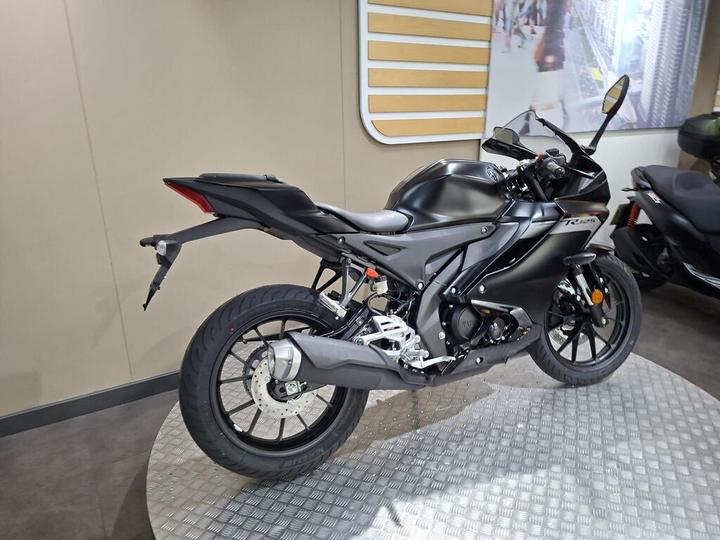 A sleek black motorcycle displayed in a showroom, showcasing its aerodynamic design and modern features, with a partial view of another motorcycle in the background.