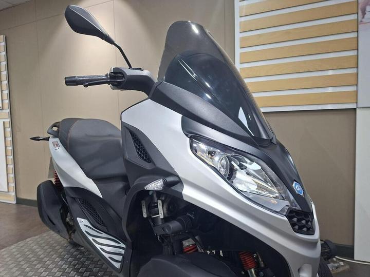 A close-up view of a silver scooter with a windscreen and black detailing, showcasing its front handlebars and headlight. The scooter is positioned in a well-lit indoor space.