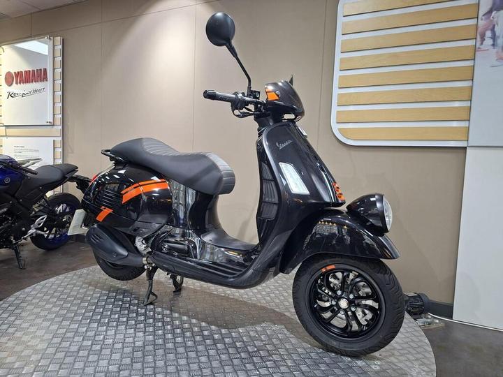 A sleek black scooter with orange accents displayed on a circular platform, set against a modern showroom backdrop featuring a Yamaha sign.