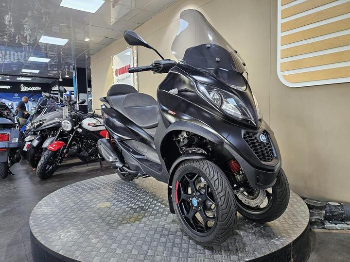 A black three-wheeled scooter displayed on a circular platform in a showroom, with other motorcycles visible in the background.