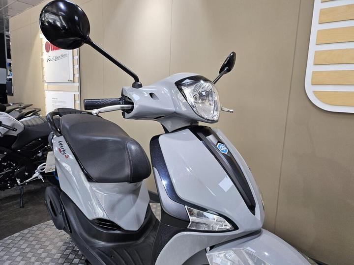 A gray scooter with a black seat and rearview mirrors, showcased against a neutral backdrop. The scooter features a prominent headlight and the model name "Liberty S" visible on the side.